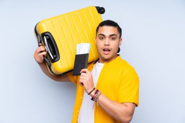 Asyalı yakışıklı adam mavi arka planda izole edilmiş, bavul ve pasaportla tatilde ve şaşırmış.