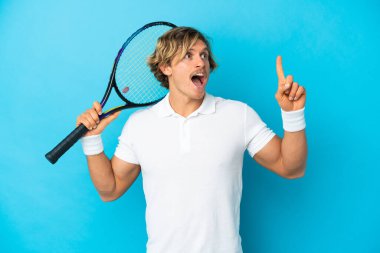 Genç sarışın adam mavi arka planda izole edilmiş tenis oynuyor ve yukarı işaret ediyor.