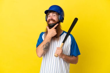 Kızıl saçlı beyzbolcu kasklı ve sopalı adam sarı arka planda izole edilmiş mutlu ve gülümsüyor.