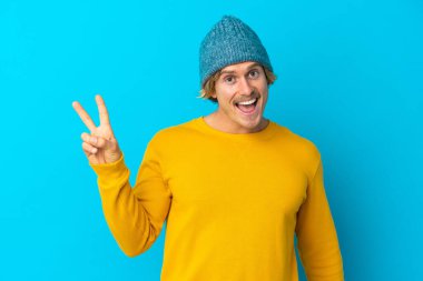 Yakışıklı sarışın adam mavi arka planda izole edilmiş gülümsüyor ve zafer işareti gösteriyor.