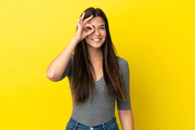 Sarı arka planda izole edilmiş genç Brezilyalı kadın parmaklarıyla tamam işareti gösteriyor