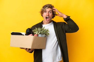 Sarışın adam, sarı arka planda izole edilmiş bir kutu dolusu şeyi alıp sürpriz bir ifadeyle hareket ediyor.