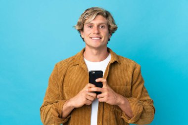 İzole edilmiş mavi arka planda İngiliz bir adam kameraya bakıyor ve cep telefonunu kullanırken gülümsüyor.