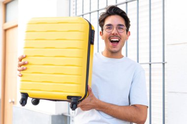 Young caucasian man at outdoors in vacation with travel suitcase and surprised