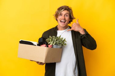 Sarışın adam, sarı arka planda izole edilmiş eşyalarla dolu bir kutuyu kaldırırken bir yandan da telefon hareketi yapıyor. Bana geri dönüş işareti ver.