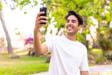 Parktaki genç beyaz adam cep telefonuyla selfie çekiyor.