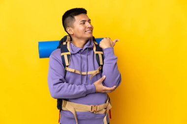 Sarı arka planda izole edilmiş büyük sırt çantalı genç dağcı bir ürünü sunmak için kenarı işaret ediyor.