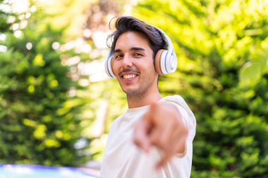 Young caucasian man in a park listening music and pointing to the front