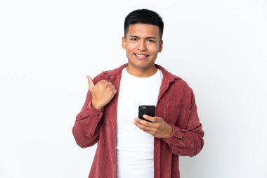 Young Ecuadorian man isolated on white background using mobile phone and pointing to the lateral