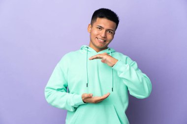Young Ecuadorian man isolated on purple background holding copyspace imaginary on the palm to insert an ad