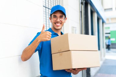 Young delivery man at outdoors holding boxes with thumb up