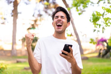 Young caucasian man in a park using mobile phone and doing victory gesture