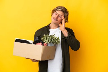 Sarışın adam, sarı arka planda izole edilmiş, baş ağrısı olan şeylerle dolu bir kutuyu kaldırırken hamle yapıyor.