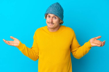 Mavi arka planda izole edilmiş yakışıklı sarışın adam ellerini kaldırırken şüphe duyuyor.