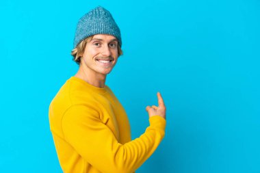 Yakışıklı sarışın adam mavi arka planda izole edilmiş geriye doğru işaret ediyor.