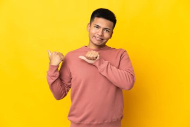 Young Ecuadorian man isolated on yellow background pointing to the side to present a product