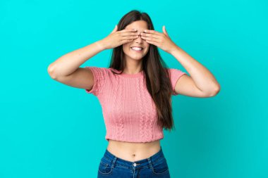 Mavi arka planda izole edilmiş genç Brezilyalı bir kadın. Gözleri elleriyle kaplıyor ve gülümsüyor.