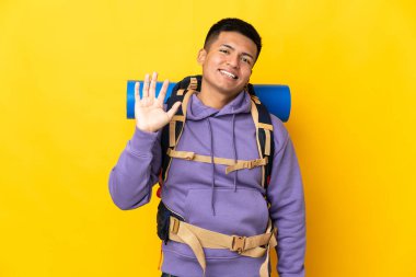 Sarı arka planda izole edilmiş büyük sırt çantalı genç dağcı mutlu bir ifadeyle selamlıyor.