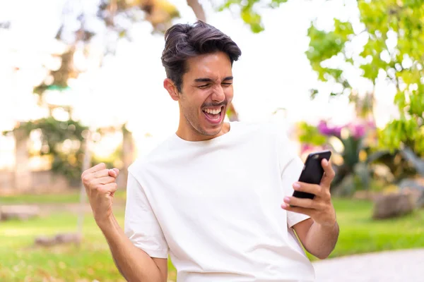 Young Caucasian Man Park Using Mobile Phone Doing Victory Gesture —  Fotos de Stock