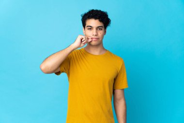 Genç Venezuelalı adam mavi arka planda izole edilmiş. Sessizlik işareti gösteriyor.
