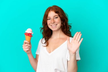 Teenager reddish woman with a cornet ice cream isolated on blue background saluting with hand with happy expression
