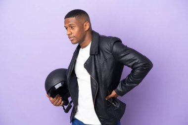 Motosiklet kasklı Latin bir adam mor arka planda izole edilmiş, gayret gösterdiği için sırt ağrısı çekiyor.