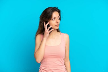 Mavi arka planda izole edilmiş cep telefonu kullanan genç beyaz kadın yan tarafa bakıyor.