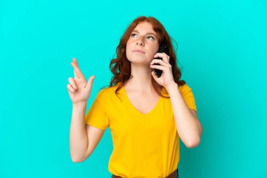 Teenager reddish woman using mobile phone isolated on blue background with fingers crossing and wishing the best