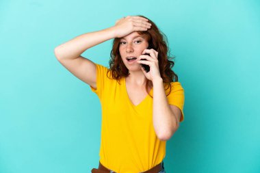 Teenager reddish woman using mobile phone isolated on blue background doing surprise gesture while looking to the side