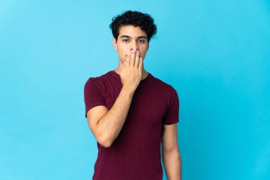 Genç Venezuelalı bir adam mavi arka planda izole edilmiş. Ağzını eliyle kapatıyor.