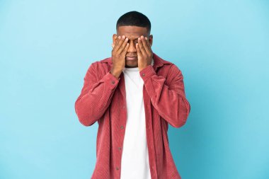 Mavi arka planda izole edilmiş genç latin adam gözlerini elleriyle kapatıyor.
