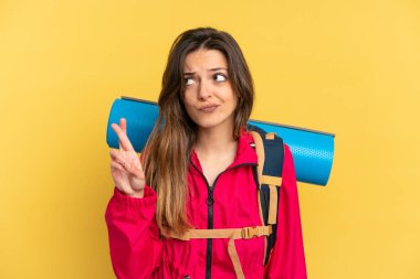 Young mountaineer man with a big backpack isolated on yellow background with fingers crossing and wishing the best