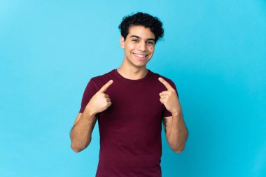 Genç Venezuelalı adam mavi arka planda izole edilmiş bir şekilde baş parmak işareti yapıyor.