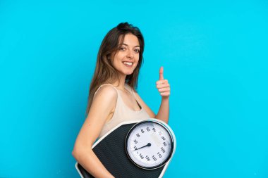 Mavi arka planda izole edilmiş beyaz bir kadın. Elinde başparmağıyla tartı makinesi tutuyor.