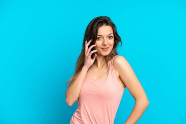 Mavi arka planda izole edilmiş cep telefonu kullanan beyaz bir kadın. Kolları kalçasında ve gülümsüyor.