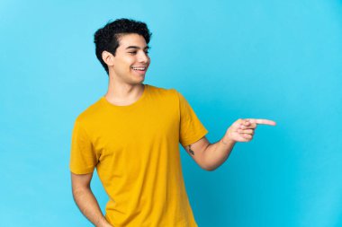 Genç Venezuelalı bir adam mavi arka planda izole edilmiş bir şekilde parmak ucunda bir ürün sunuyor.