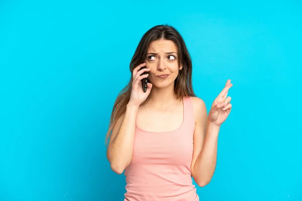 Молодая Кавказская Женщина Используя Мобильный Телефон Синем Фоне Скрестив Пальцы — стоковое фото