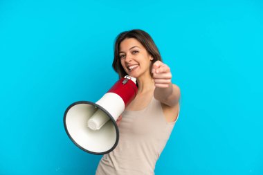 Mavi arka planda izole edilmiş genç beyaz kadın elinde bir megafon, elinde gülümseyerek ön tarafı işaret ediyor.