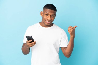 Genç latin adam mavi arka planda cep telefonu kullanarak ve yan tarafı işaret ederek izole edildi