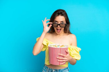 Mavi arka planda soyutlanmış beyaz bir kadın 3D gözlüklü ve elinde koca bir kova patlamış mısırla şaşırdı.