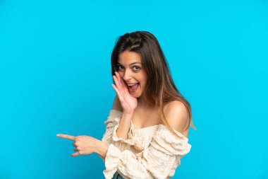 Mavi arka planda izole edilmiş genç beyaz kadın bir ürünü sunmak ve bir şeyler fısıldamak için kenarı işaret ediyor.