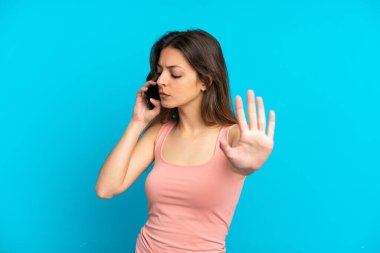 Mavi arka planda izole edilmiş cep telefonu kullanan beyaz bir kadın dur hareketi yapıyor ve hayal kırıklığına uğruyor.