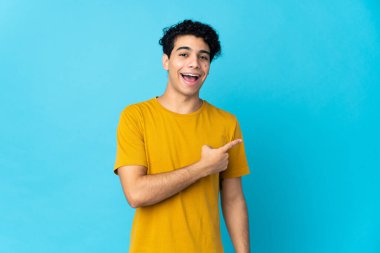 Genç Venezuelalı adam mavi arka planda izole edilmiş, şaşırmış ve işaret ediyor.