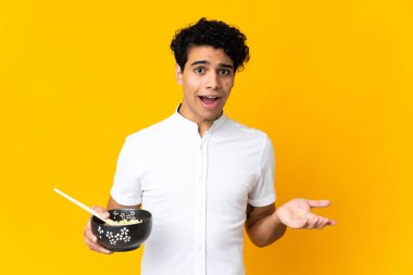 Genç Venezuelalı bir adam, elinde yemek çubuklarıyla bir kase erişte tutarken, şok olmuş yüz ifadesiyle sarı arka planda izole edilmiş.