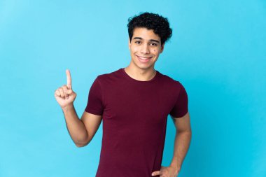 Genç Venezuelalı adam mavi arka planda izole edilmiş ve en iyinin işareti olarak parmağını kaldırıyor.