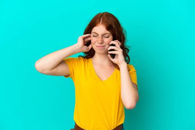 Teenager reddish woman using mobile phone isolated on blue background frustrated and covering ears
