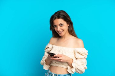 Mavi arka planda izole edilmiş genç beyaz kadın cep telefonuyla bir mesaj gönderiyor.