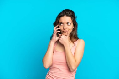 Mavi arka planda izole edilmiş cep telefonu kullanan beyaz bir kadın. Şüpheleri ve düşünceleri var.
