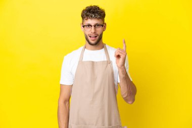 Restoran garsonu. Sarışın adam sarı arka planda izole edilmiş. Parmakla işaret eden bir fikir düşünüyor.