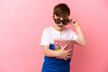 Pembe arka planda izole edilmiş küçük kızıl saçlı çocuk 3D gözlüklü ve elinde koca bir kova patlamış mısırla şaşırdı.